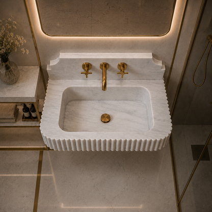 A modern wall-mounted sink with a fluted white marble-look front and basin, paired with brushed gold faucet hardware, shown in a styled bathroom setting with warm lighting.