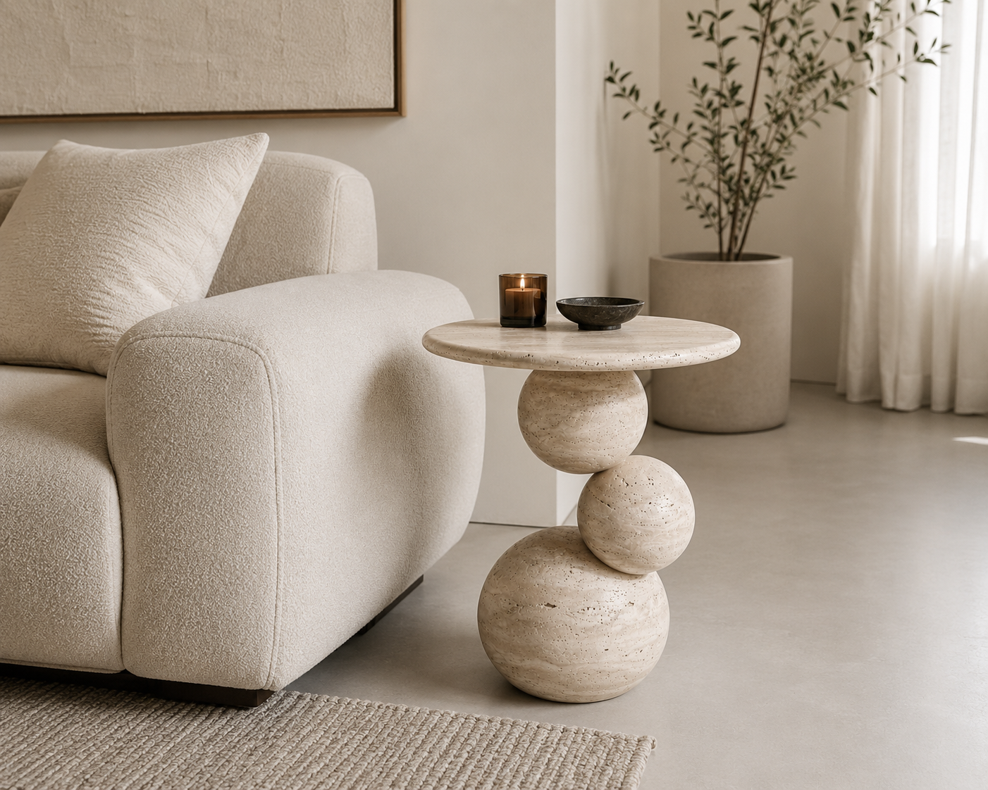 A round stone side table with a sculptural stacked-sphere base, styled in a minimalist living room next to a cream bouclé sofa.