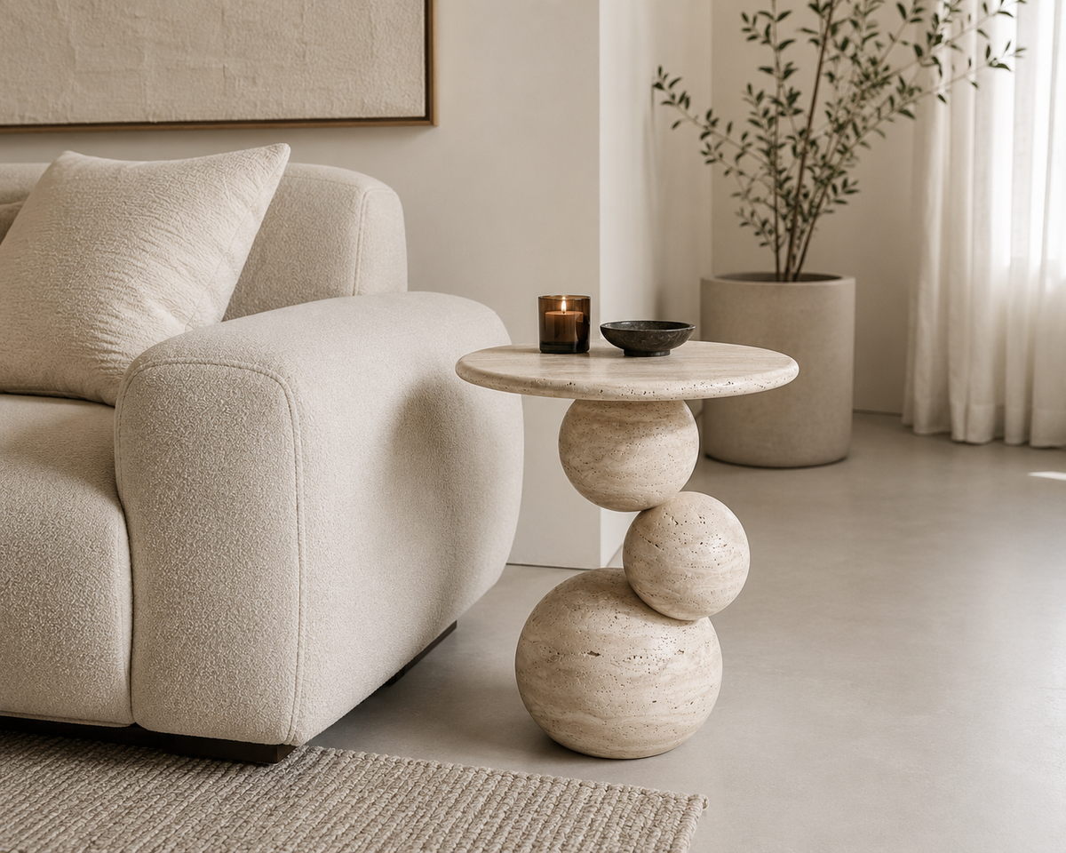 A round stone side table with a sculptural stacked-sphere base, styled in a minimalist living room next to a cream bouclé sofa.