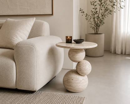 A round stone side table with a sculptural stacked-sphere base, styled in a minimalist living room next to a cream bouclé sofa.