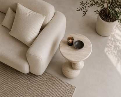 A styled interior scene showing a cream upholstered sofa with matching pillows, a round stone side table with a candle and bowl, a potted plant, and a woven rug on a light concrete floor.